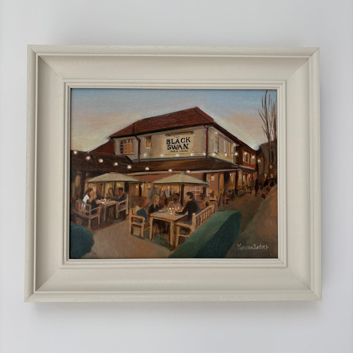 Framed oil painting of The Black Swan pub by Minuca Sostres, showing diners outside beneath parasols and glowing string lights at dusk.