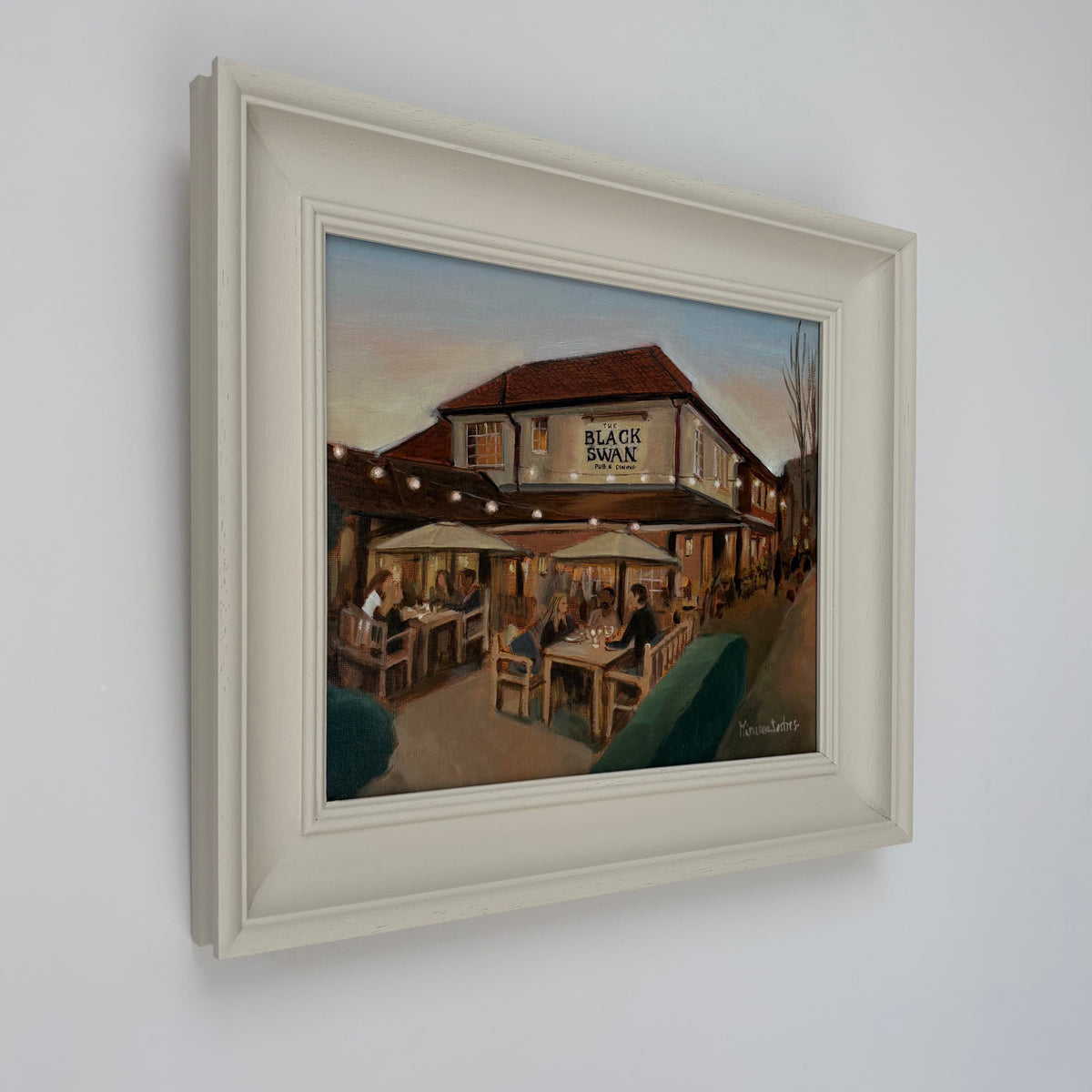 Framed oil painting of The Black Swan pub by Minuca Sostres, showing diners outside beneath parasols and glowing string lights at dusk.